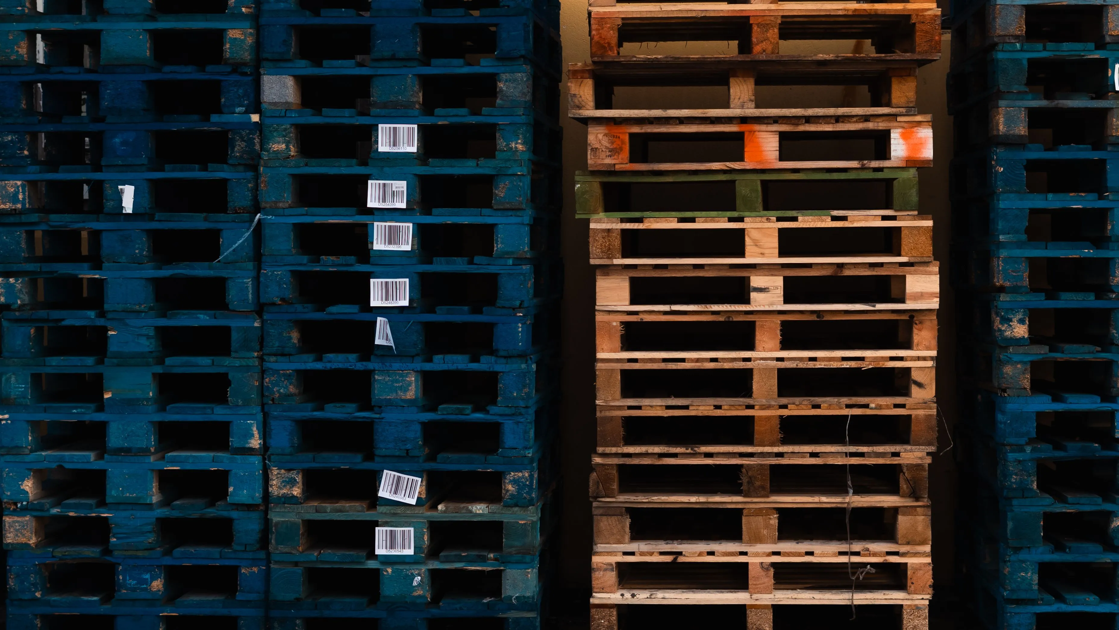 Tall stacks of wooden shipping pallets with barcode labels in a freight warehouse, lit in warm and cool graphite tones.
