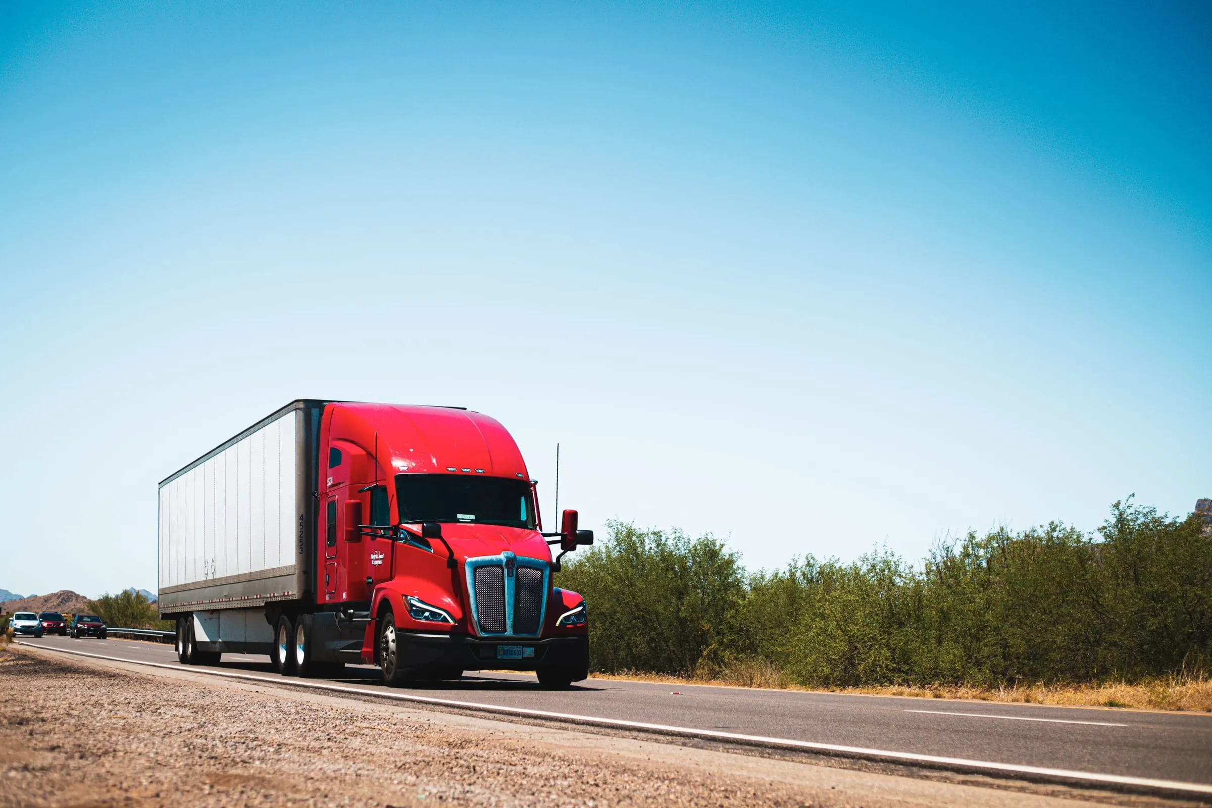 A long-nose semi tractor pulling a freight trailer down an open highway under a wide sky, in graphite and warm light.